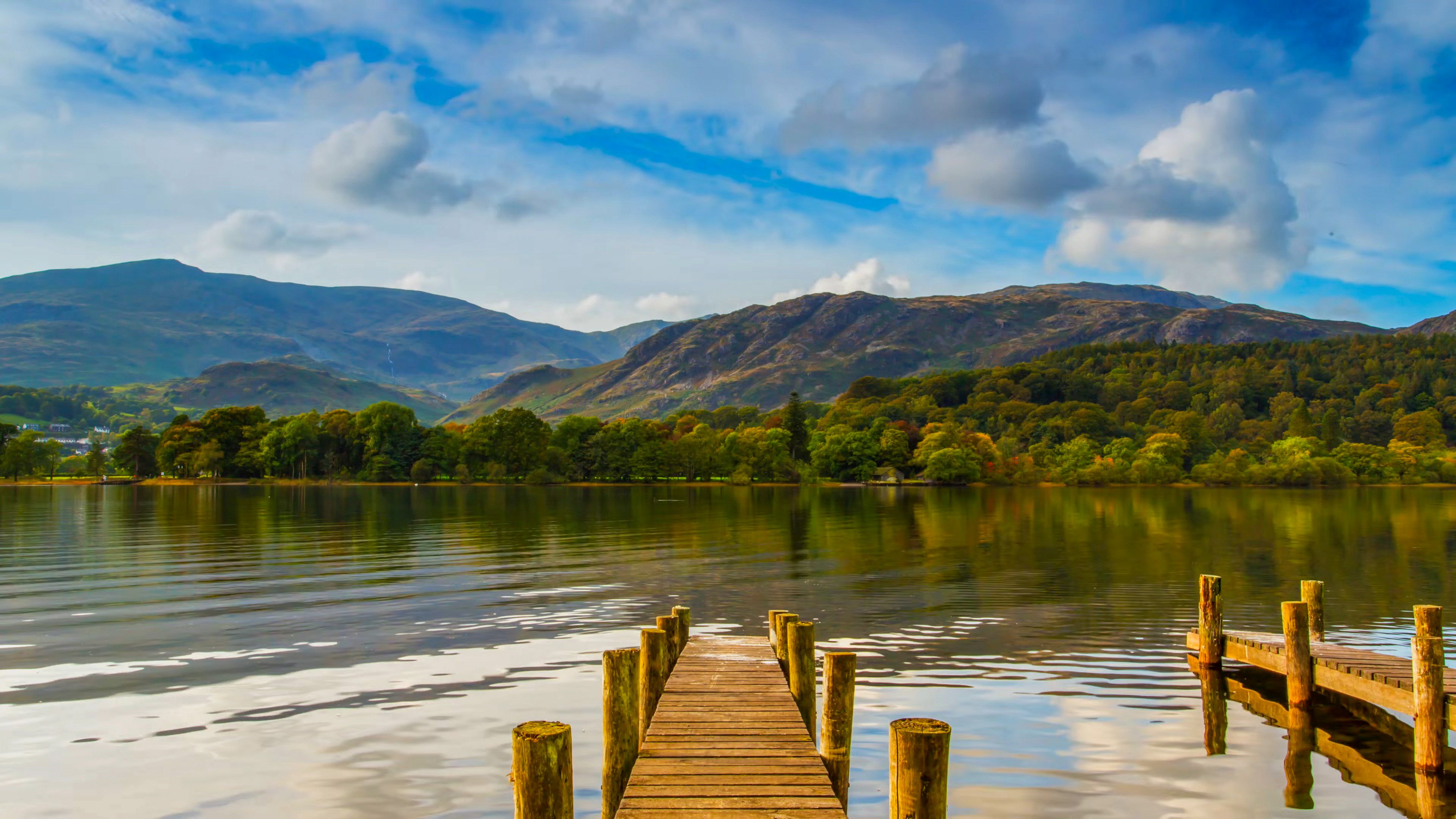 A wooden pier extends into a calm lake, reflecting the surrounding green hills and distant mountains under a partly cloudy blue sky.