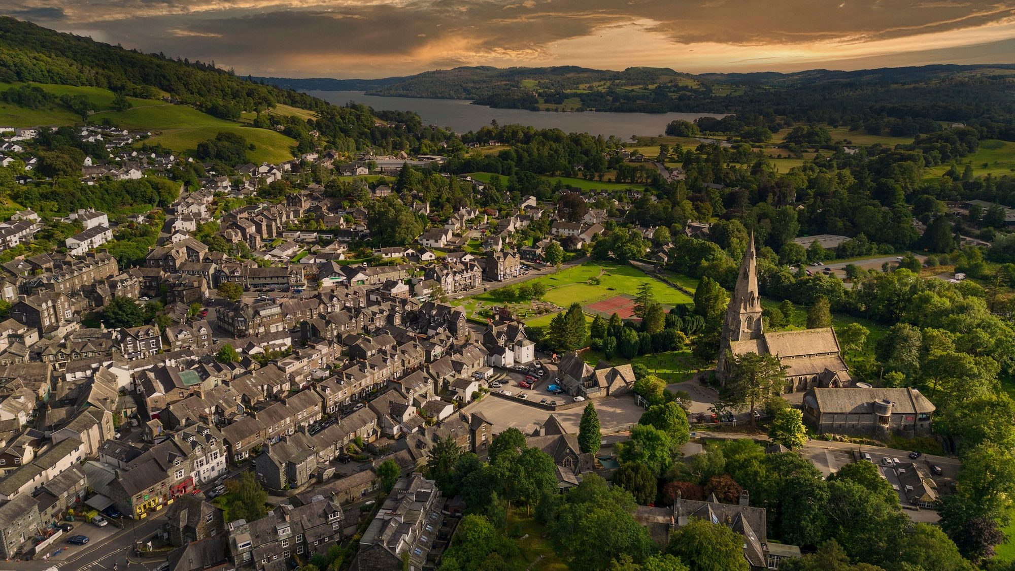 Aerial view of Ambleside in the Lake District, showing stone buildings, a church with a tall spire, green hills, and Lake Windermere at sunset.