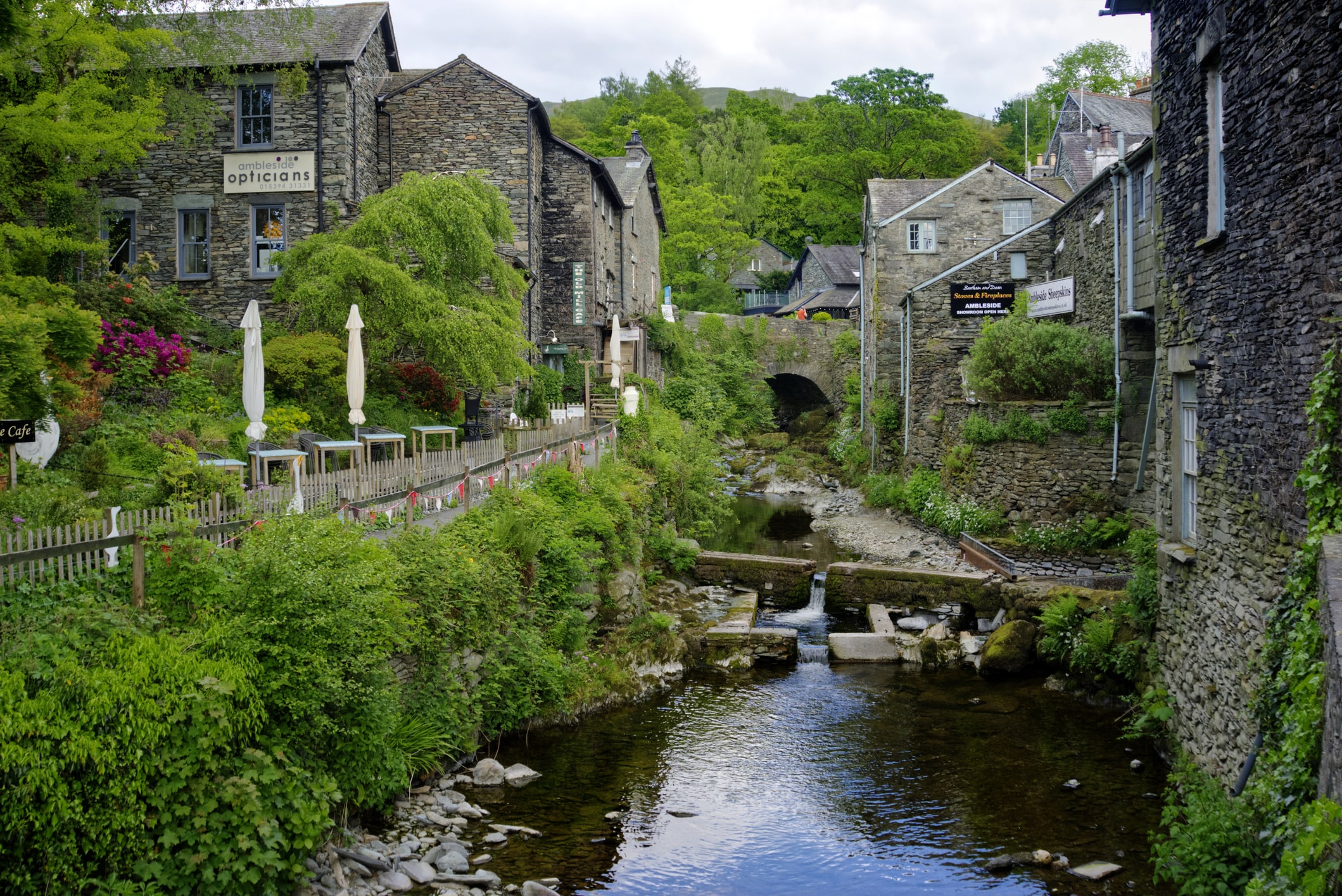 A narrow stream runs between old stone buildings and shops in Ambleside, Lake District, with a garden café, greenery, and a small arched bridge ahead.