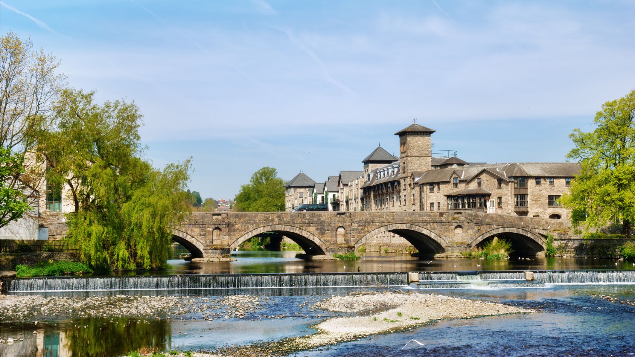 Stone bridge over River Kent in Kendal, Cumbria, with historic buildings, a small weir, and lush trees under a bright blue sky.