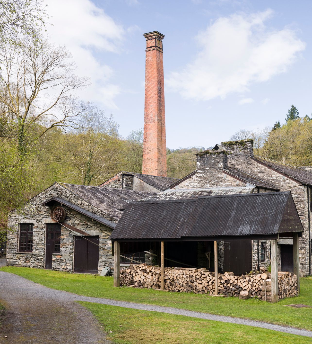 A historic stone building with a tall red brick chimney stands in a wooded area. Logs are stacked under a covered shelter, and a path winds through the grassy yard.