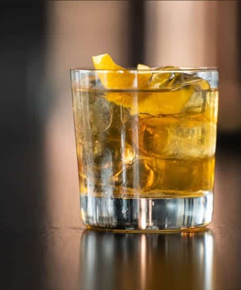 Cask-aged whiskey on a dark surface with ice and lemon garnish.